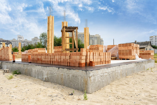 Angle View Rows Of New Red Brick Wall Lies On Foundation Of House Under Construction. Residential Building With Height Marks Install Wooden Support Element And Concrete Column Formwork