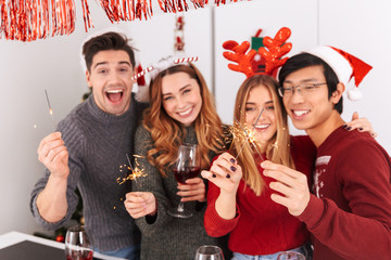 Group of happy multiethnic people holding glasses with wine and sparklers, while celebrating New Year in flat