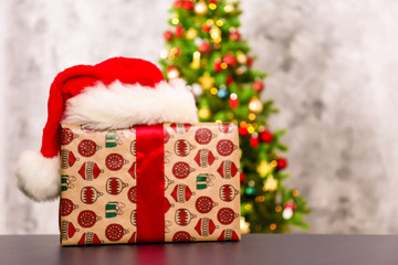 Festive composition with one single present in colorful wrapping paper with santa claus hat on foreground and blurry decorated Christmas spruce tree on background. Close up, copy space.