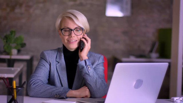 Portrait of middle-aged blonde short-haired businesswoman in glasses gladly talking on cellphone in office.
