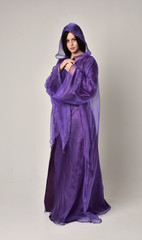 Naklejka premium full length portrait of beautiful girl with long black hair, wearing purple fantasy medieval gown and cloak. standing pose on grey studio background.