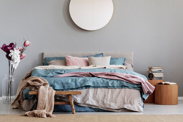 Trendy round mirror on grey wall in chic bedroom interior with cozy bed with blue, pastel pink and beige sheets and wooden nightstands with books
