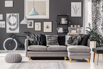 Knot pillow on scandinavian grey couch in monochromatic living room interior with pouf, bike and posters on the wall
