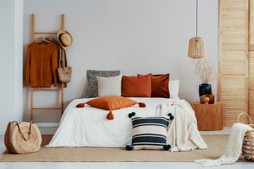 Copy space on white empty wall of trendy bedroom with flower in vase on wooden nightstand, king size bed with autumn colored bedding and wooden ladder - obrazy, fototapety, plakaty