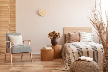 Stylish retro armchair with white pillow next to two round wooden tables with books and vase with flowers in beige bedroom interior