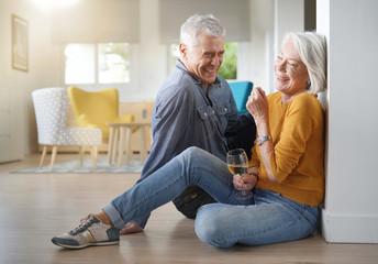 Relaxed senior couple sitting on floor in modern home with glass of wine - Powered by Adobe