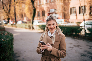 Fototapeta premium Portrait of a smiling young woman in coat using smart phone outdoors.