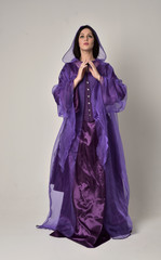 Naklejka premium full length portrait of beautiful girl with long black hair, wearing purple fantasy medieval gown and cloak. standing pose on grey studio background.