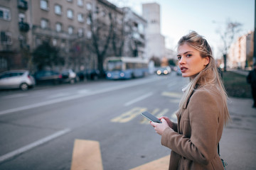 Beautiful young stylish woman using smartpone while waiting for taxi or bus in the city.