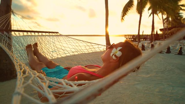 SLOW MOTION, CLOSE UP, LENS FLARE: Relaxed woman swinging on the beach while lying in a hammock on a beautiful summer evening. Carefree girl sleeping in a hammock at sunset in scenic Seychelles.