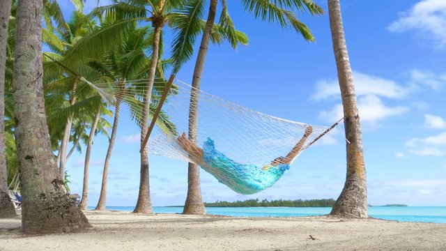 LOW ANGLE: Female traveler unwinds by the breathtaking ocean by taking an afternoon nap in a rope hammock. Relaxed woman enjoying her summer vacation on tropical island by swaying under the palm trees