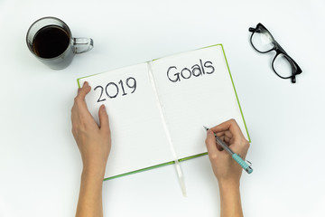 New Year, new me concept. Top view of female hands writing "2019 goals" in a copy book on white table
