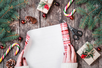 Female hand wrap Christmas gift in holiday paper on wooden background with Fir branches, candy, decorations. Happy New Year