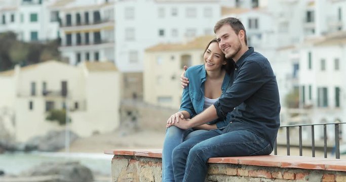 Happy couple of tourists contemplating ocean sitting on a ledge in a coast town on vacation