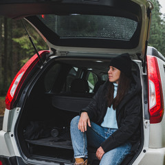 young adult woman sitting in suv trunk resting after road trip at road side in mountain forest