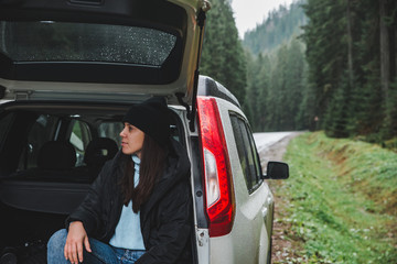 young adult woman sitting in suv trunk resting after road trip at road side in mountain forest