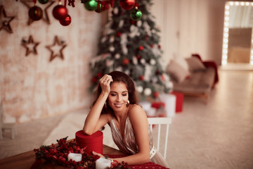 Winter holidays decorations. Warm colors. Charming brunette woman in beige dress sits at the red table and holds a round present box before a Christmas tree