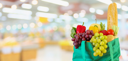 Fresh fruits and vegetables in reusable green shopping bag with supermarket grocery store blurred defocused background with bokeh light