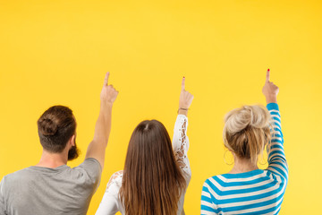 Back view of modern young people standing in row on yellow backdrop showing up with fingers