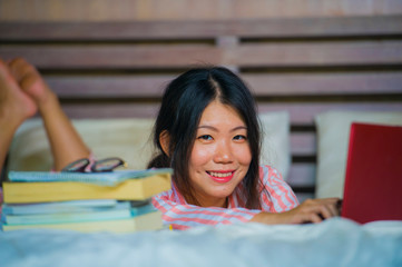 young beautiful and smart Asian Korean student woman at home bedroom sitting on bed studying for exam with laptop computer and textbooks smiling confident