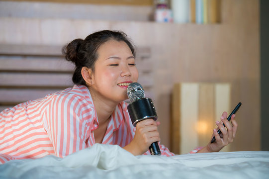 Beautiful Asian American Teenager Girl Singing Karaoke Song Excited At Home Bedroom Holding Mobile Phone Playing On Bed Excited And Cheerful Emulating Pop Music Star