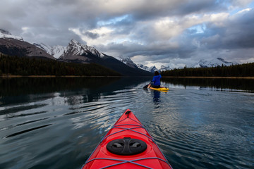 Kayaking in a peaceful and calm glacier lake during a vibrant cloudy sunset. Taken in Maligne Lake, Jasper National Park, Alberta, Canada.