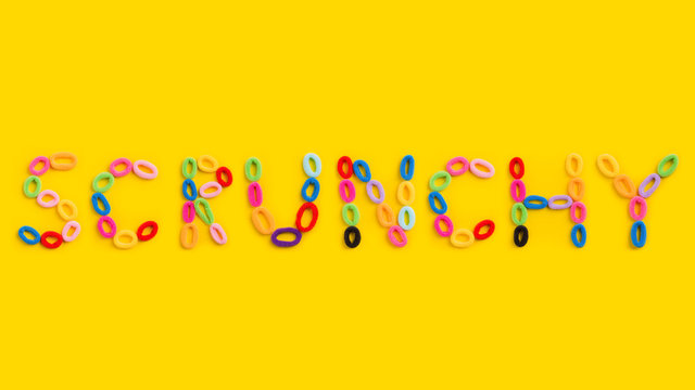 The word "Scrunchy" laid out of colored rubber bands on a yellow background. Colored hair ties on a yellow background.