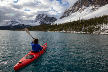 Adventurous girl kayaking in a glacier lake surrounded by the Canadian Rockies during a cloudy morning. Taken at Bow Lake, Banff, Alberta, Canada.