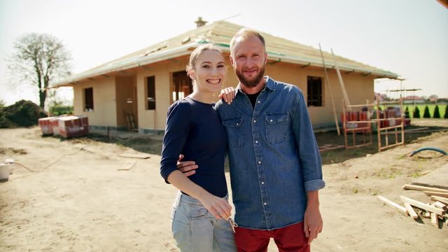 Two young people standing outdoors on the construction site in front of their new house. Medium shot. Soft focus