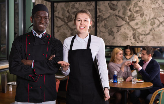 Polite waitress standing in restaurant hall with cheerful African American chef, meeting guests