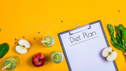 The diet plan is written on a white sheet on the tablet among the bottles of green smoothies, spinach and apples on a bright yellow background. Slimming and detox concept