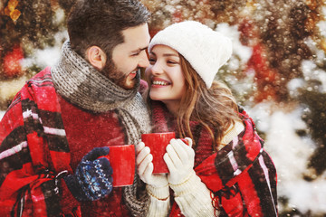 Young beautiful happy smiling couple holding red cups, mugs of hot drink. Models looking at each other, bundled up tartan blanket. Winter holidays, Christmas celebration, winter holidays concept
