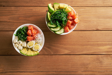 Raw Organic Poke Bowl with with rice, avocado, salmon, mango, cucumbers, chuka salad, quail eggs, sweet onions two plates close-up on wooden background top view