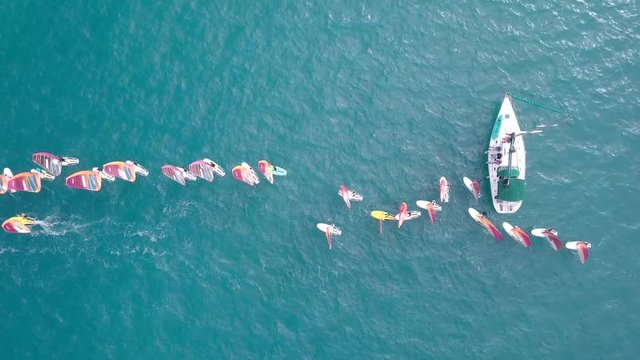 Small white yacht surrounded by a large group of Wind surfers at The Mediterranean Sea - Top down aerial footage.