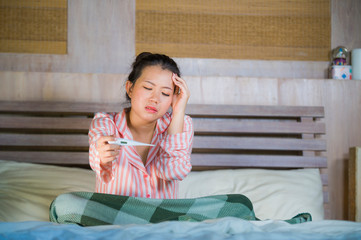 young beautiful and sweet Asian Korean woman in pajamas feeling sick at home bedroom suffering cold and flu taking temperature with thermometer in bed
