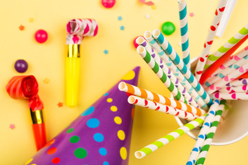 bright birthday party background with cocktail tubes, hats, horn and candy. multicolored holiday accessories on yellow table. top view