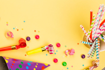 bright birthday party background with cocktail tubes, hats, horn and candy. multicolored holiday accessories on yellow table. top view