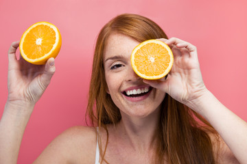 Funny young woman holding orange slices at her face and looking at camera over pink background.