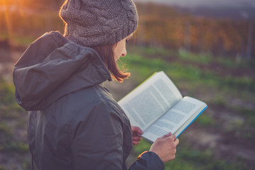 girl with jacket and hoodie reading book at park in autumn sunset light