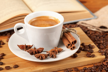 Coffee break a Cup of hot coffee and a book on a wooden table. A white Cup of black coffee surrounded by a small amount of roasted coffee beans, cinnamon, anise and an open book