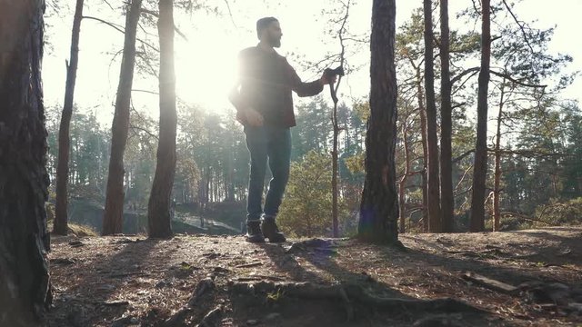 Young bearded man with an axe walk in forest and cuting a tree. Unshaven forester with an ax outdoors.