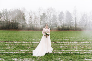 A beautiful portrait of the bride in a stylish dress with a winter bouquet of roses, cotton and spruce against a green field with the first . Wedding photo