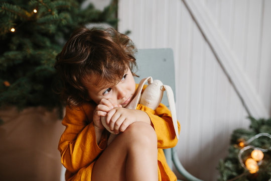 Cute curly toddler girl wearing mustard linen dress in studio near christmas tree.