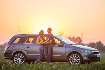 Obraz na płótnie Canvas Young couple, slim attractive woman with long hair and handsome sportive man stand embraced together at silver car on warm summer evening on bright sky at sunset or sunrise copy space background.
