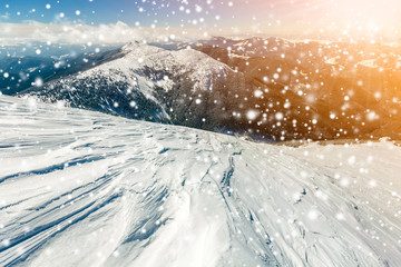 Beautiful winter landscape. Steep mountain hill slope with white deep snow, distant mountain range panorama, large snowflakes and bright shining sun rays on blue sky colorful copy space background.