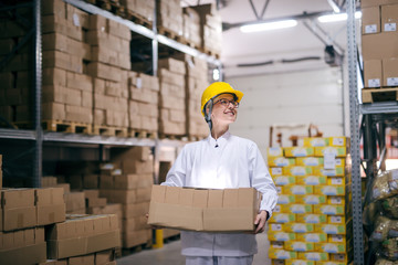 Smiling female worker carrying boxes in warehouse. Protective helmet on head, all around boxes on...