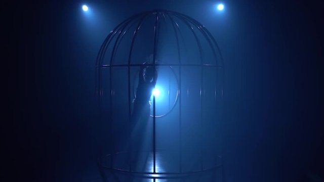 Girl in a cage stunts on a hoop in a dark room. Blue smoke background. Silhouette. Slow motion