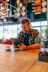 Young Smiling Caucasian bearded freelancer using tablet for video call while sitting in cafeteria. Headphones on ears and coffee on the table.