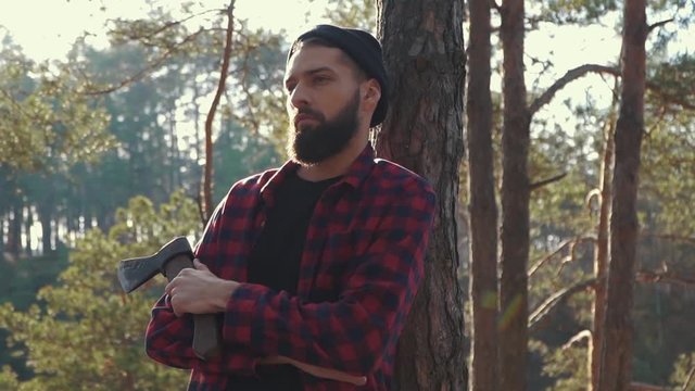Brutal serious man stand near tree with axe in hand, thinking.
