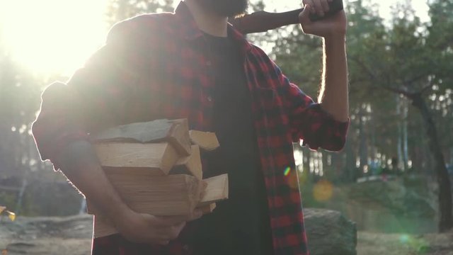 Bearded guy stand in the forest with chopped wood and axe in hands. Forester carries firewood.